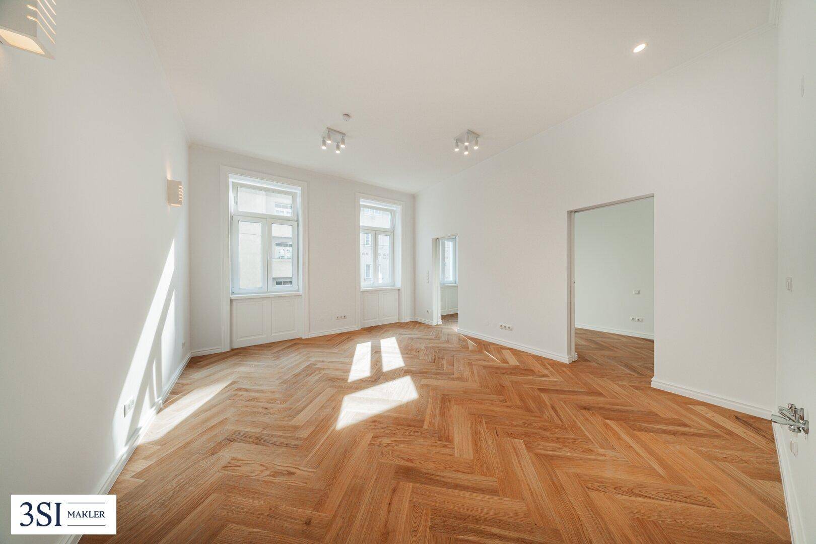 Geräumiges Zimmer mit elegantem Fischgrätparkett, weißen Wänden und Zugang zu einem weiteren hellen Raum.