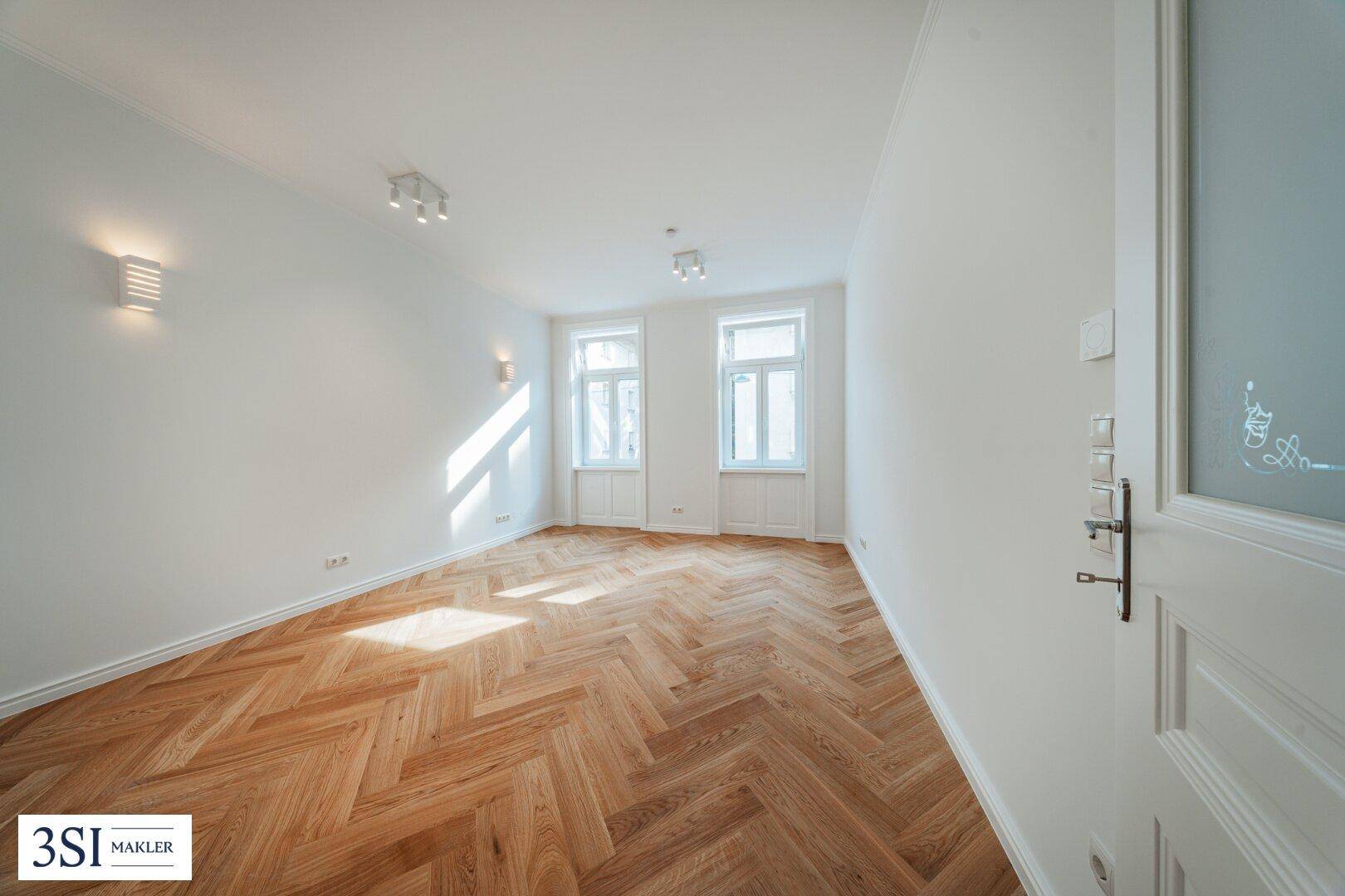 Geräumiger, heller Raum mit Fischgrätparkett und zwei hohen Fenstern, ideal für Wohn- oder Schlafzimmer.