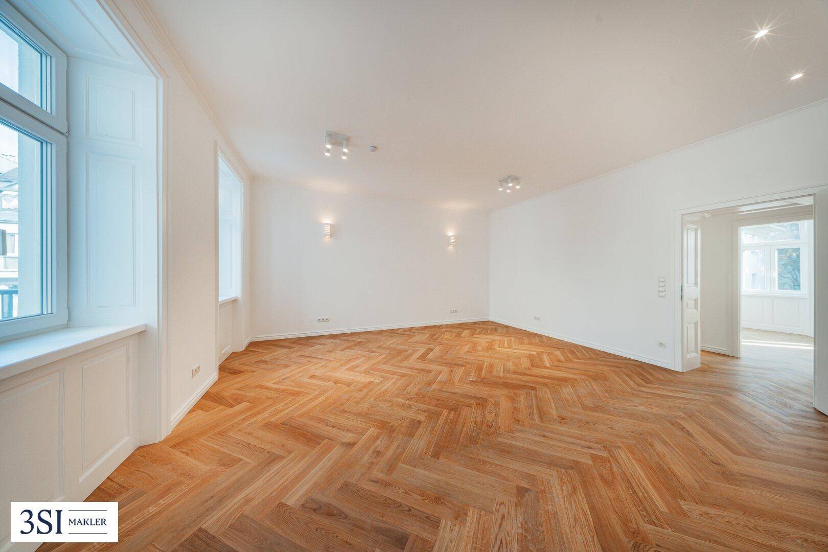 Geräumiges Wohnzimmer mit Fischgrätparkett, hohen Decken und viel Tageslicht durch mehrere Fenster.