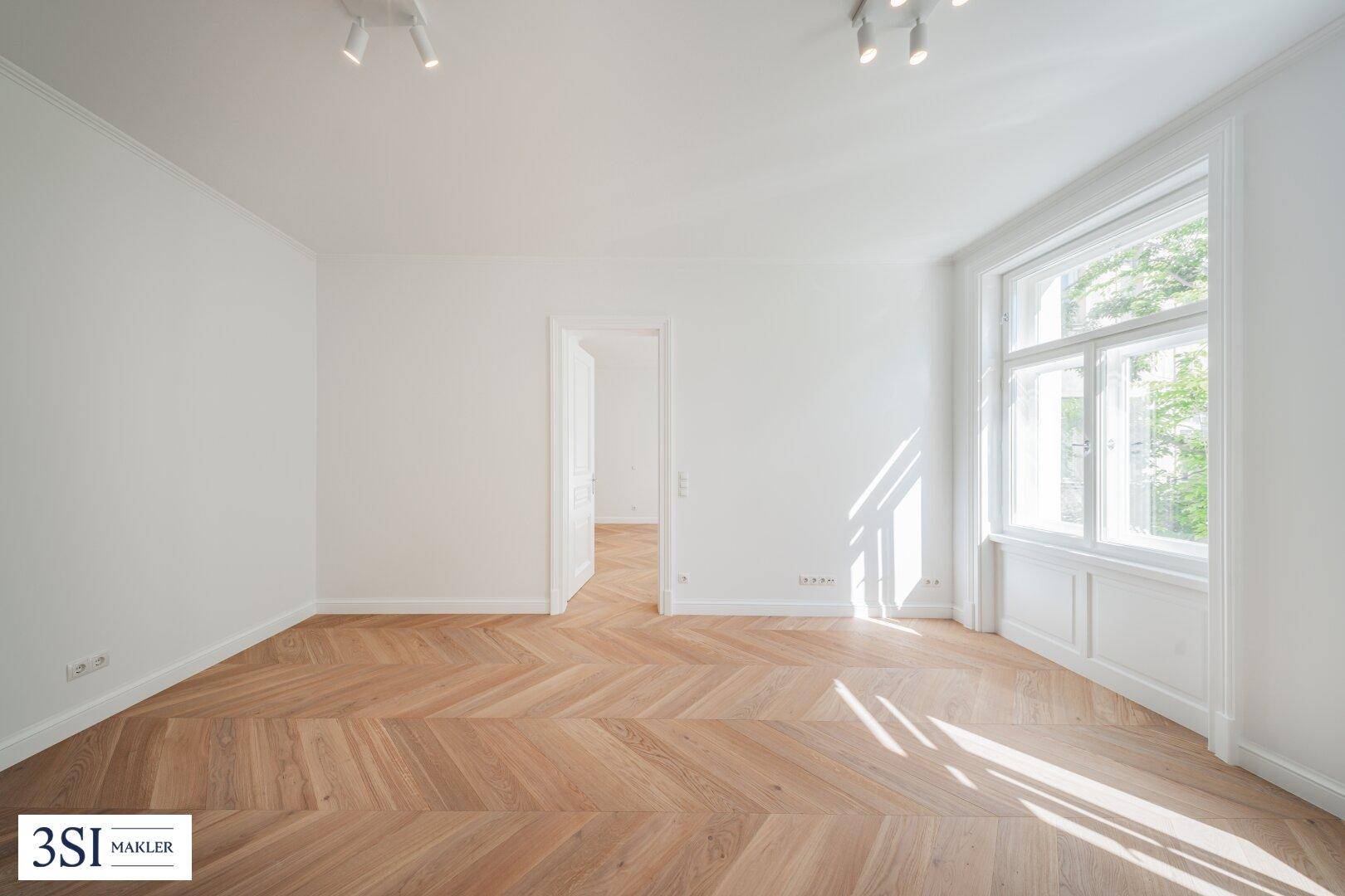 Lichtdurchflutetes Zimmer mit klassischem Fischgrätparkett und klaren, weißen Wänden, ideal für ein Wohnzimmer.