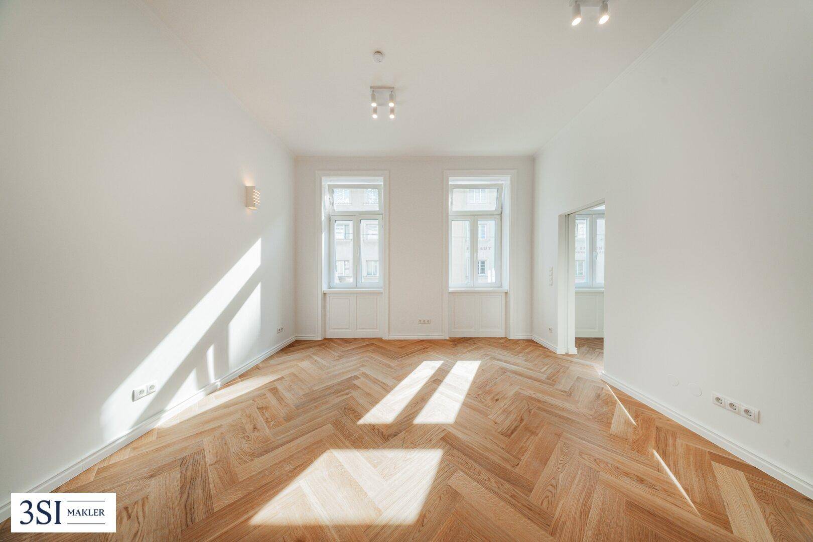 Lichtdurchfluteter Raum mit elegantem Fischgrätparkett und zwei großen Fenstern, die viel Sonne hereinlassen.