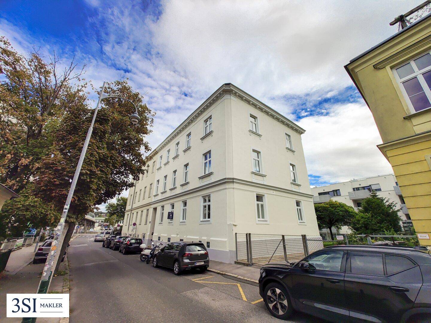 Eckgebäude an einer Straße mit parkenden Autos und Bäumen unter blauem Himmel.