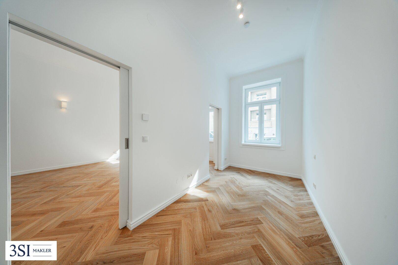 Heller Innenraum mit Fischgrätparkett, einem Fenster und offenem Durchgang zu einem weiteren Zimmer.