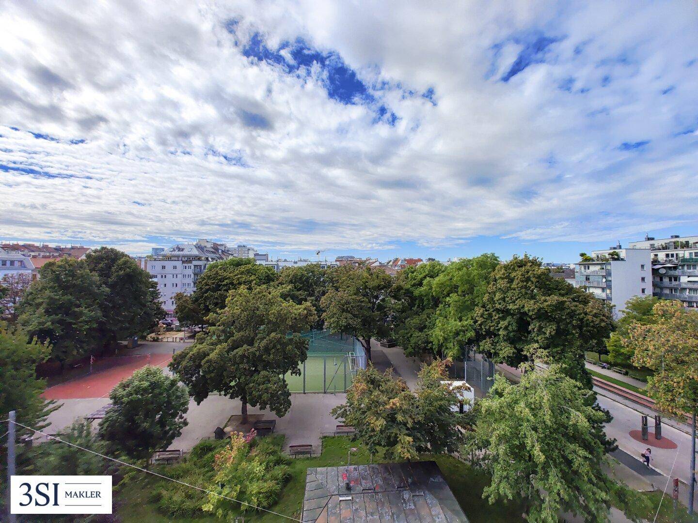 Panoramablick über eine grüne Parkanlage mit Sportplatz und umliegenden Stadtgebäuden unter bewölktem Himmel.
