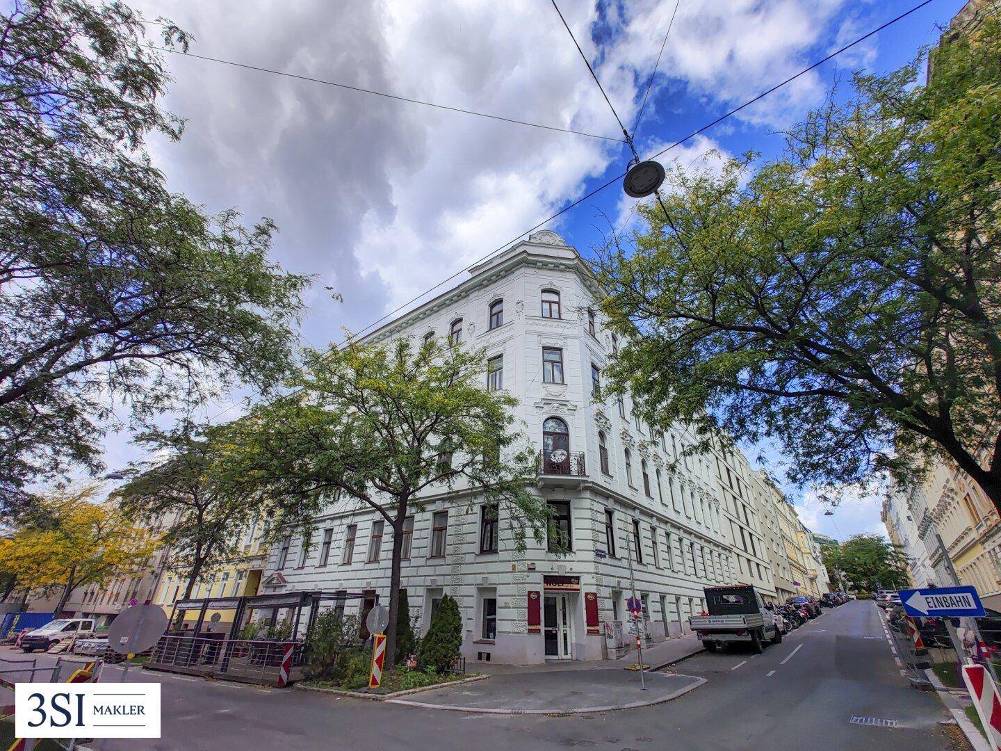 Historisches Eckgebäude an einer belebten Straße mit Bäumen und parkenden Autos.