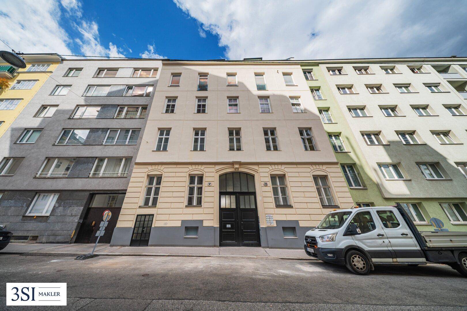 Fassade eines Mehrfamilienhauses an einer Straße mit parkenden Fahrzeugen.