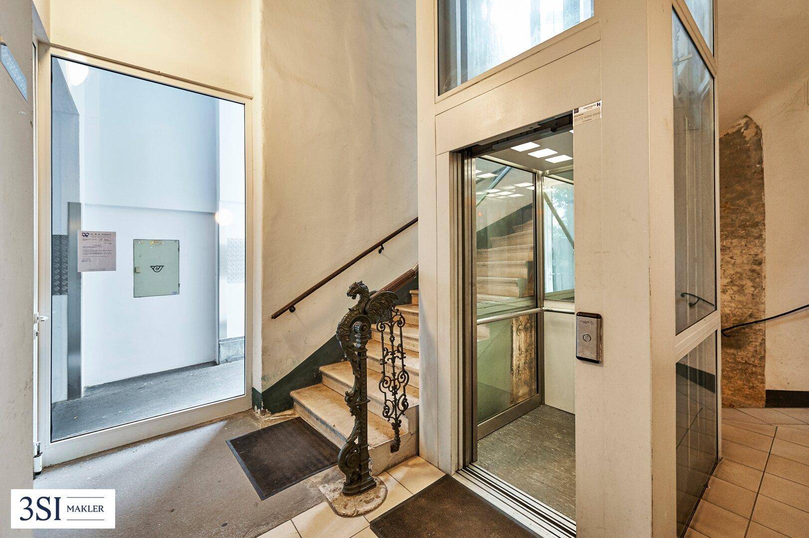 Heller Eingangsbereich mit Treppe und modernem Glasaufzug im historischen Gebäude.