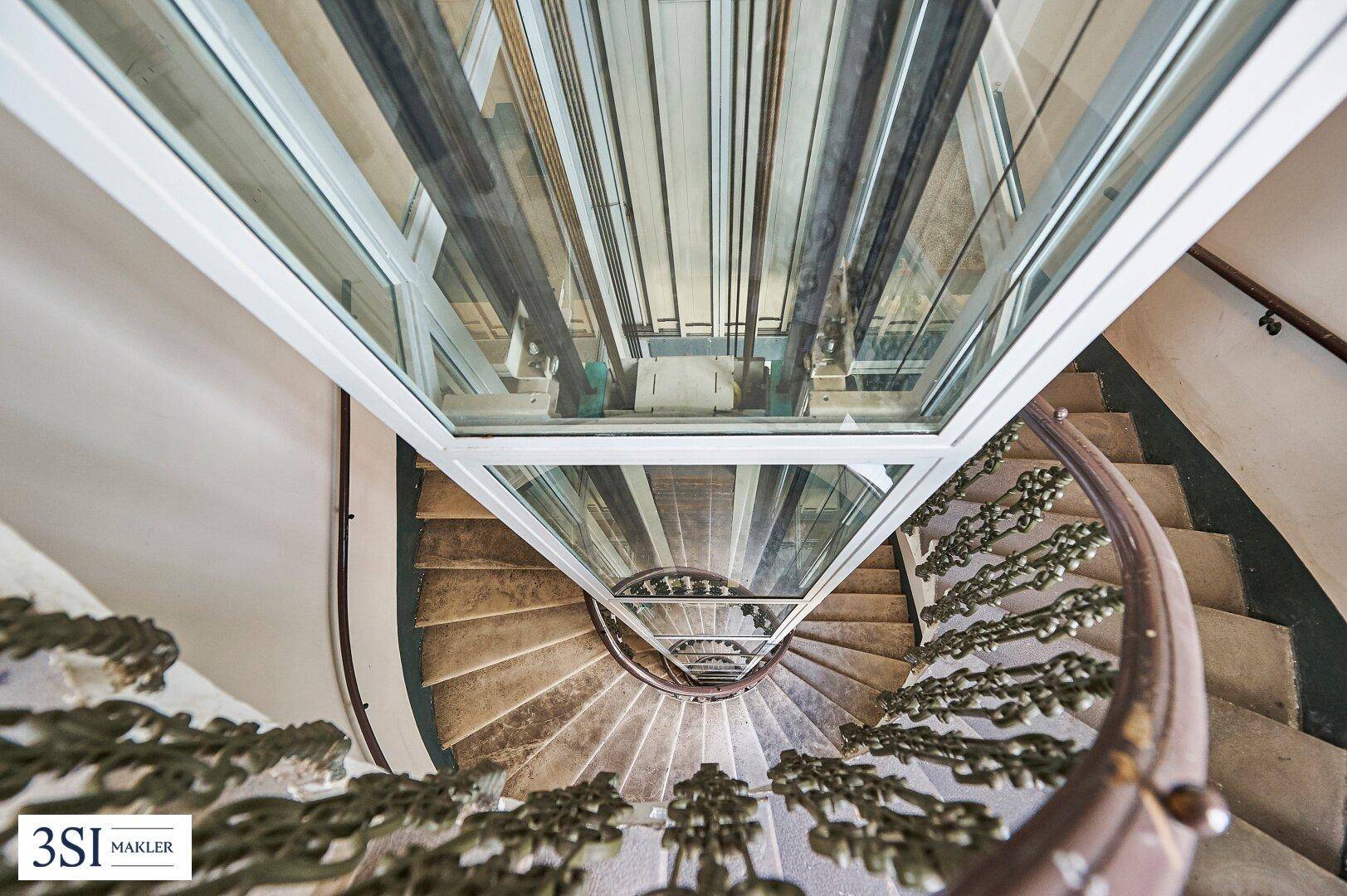 Blick von oben auf eine Wendeltreppe und den gläsernen Aufzugschacht im Treppenhaus.