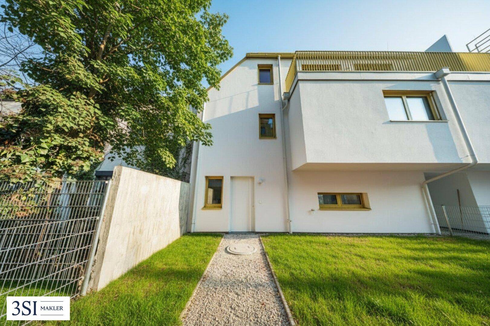 Rückseite eines modernen Gebäudes mit kleinem Rasen, Kiesweg und Bäumen.