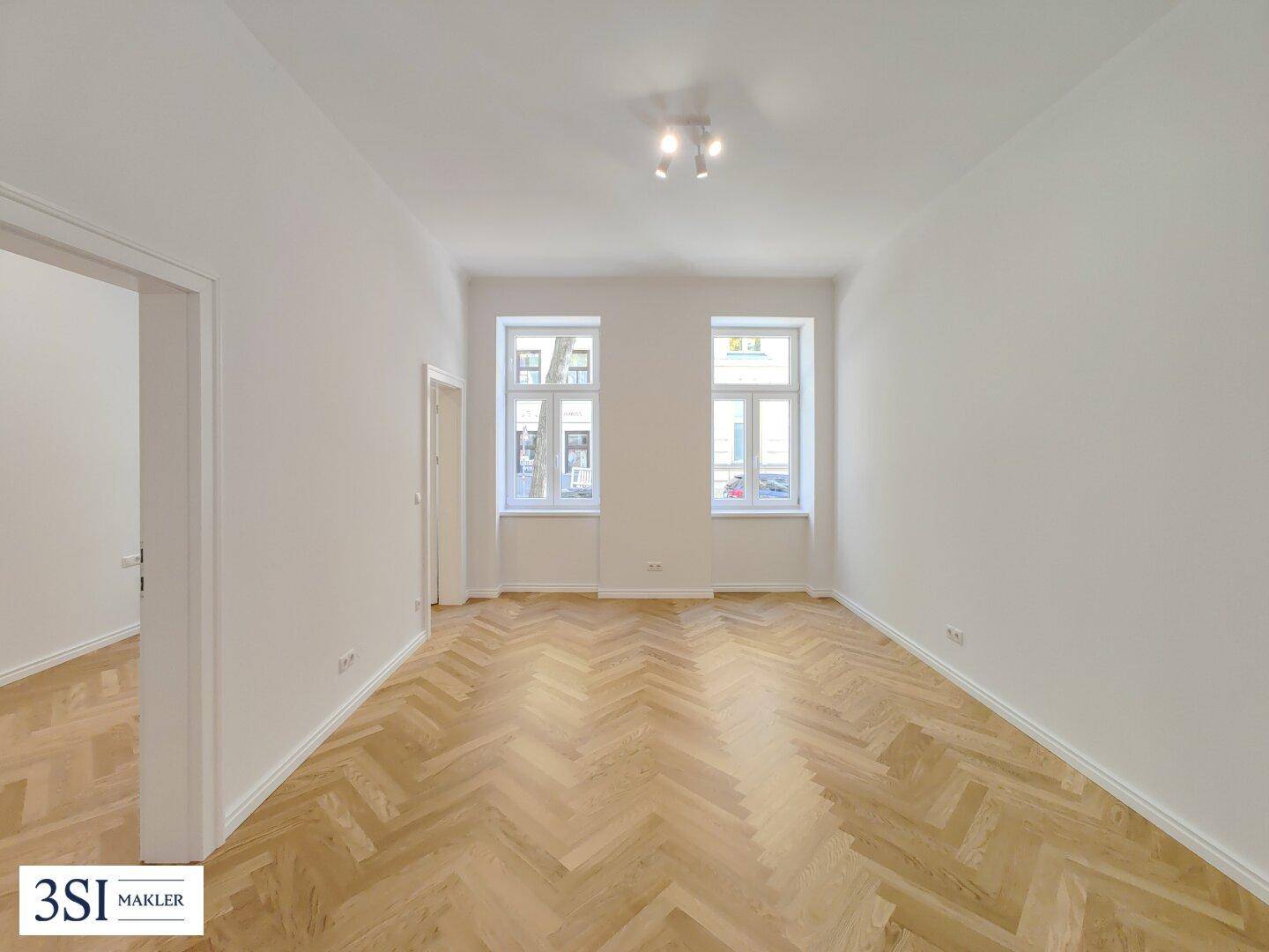 Helles, leeres Zimmer mit Fischgrätparkett und zwei Fenstern, ideal für Wohn- oder Schlafbereich.