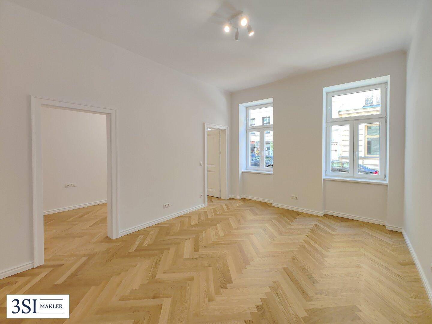Geräumiges, leeres Zimmer mit hellem Fischgrätparkett und zwei großen Fenstern, bereit zur Einrichtung.