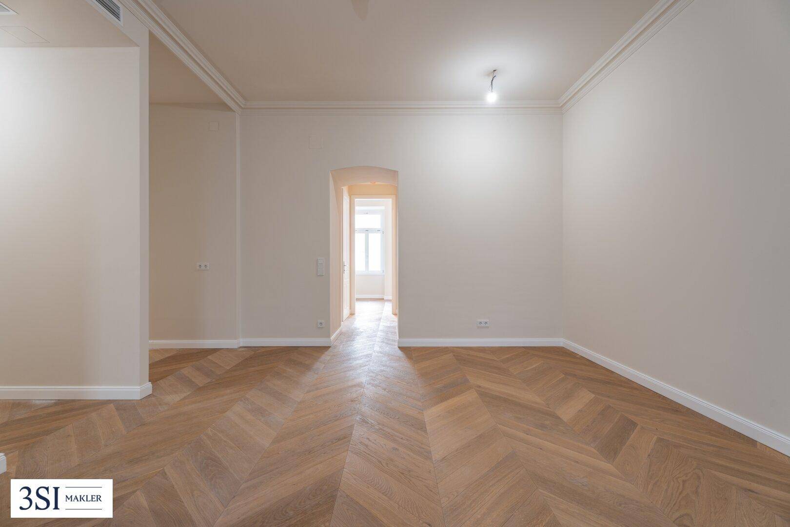 Großzügiger Raum mit Fischgrätparkett und einem eleganten Durchgang zu einem weiteren Zimmer.