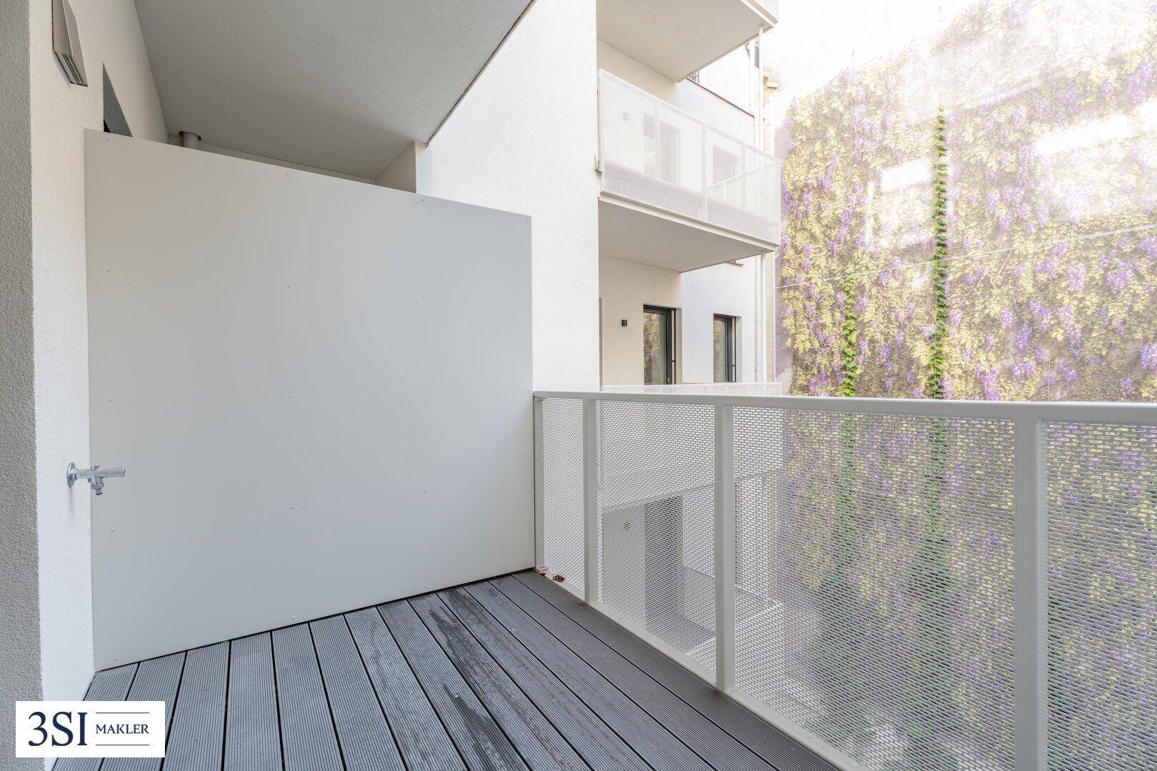 Heller Balkon mit modernen Dielen und weißem Geländer, umgeben von grüner Bepflanzung.