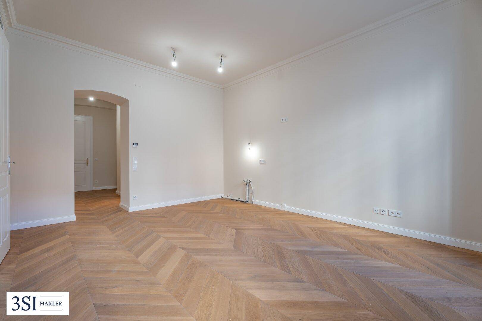 Geräumiges Zimmer mit Fischgrätparkettboden, hohen Decken und einem Bogenzugang.