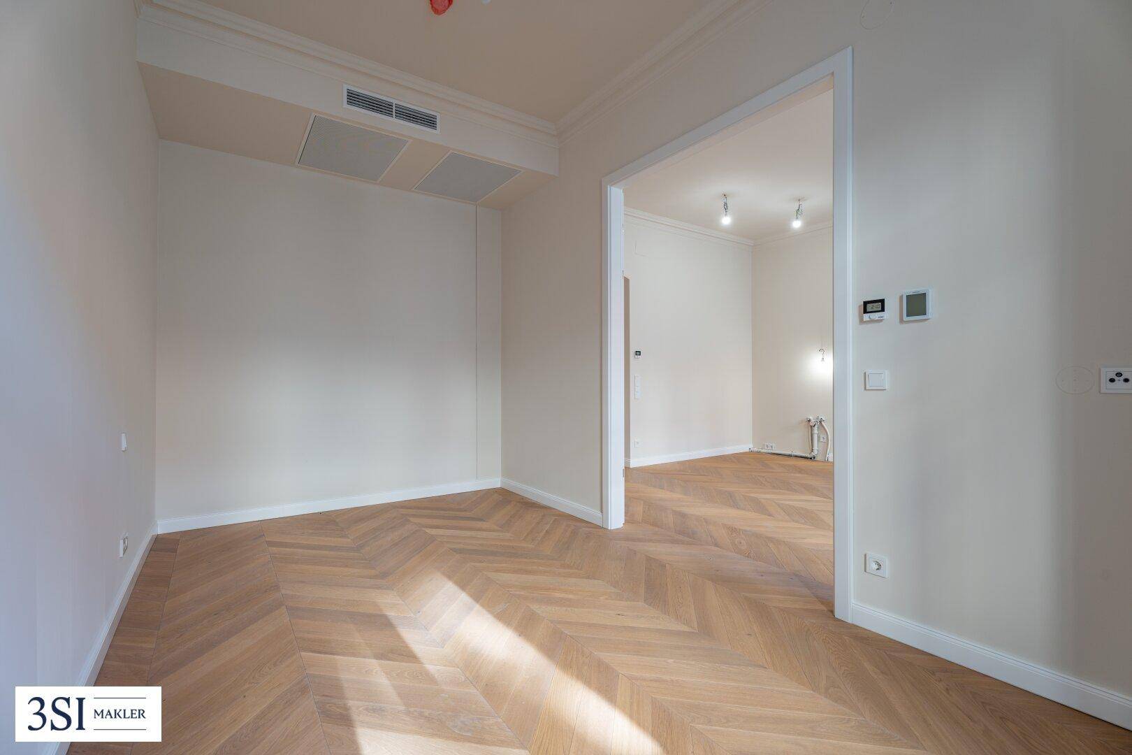 Heller Raum mit Fischgrätparkett und Zugang zu einem weiteren Zimmer mit Klimaanlage.