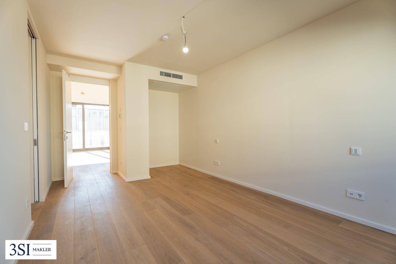 Leeres Zimmer mit Holzboden und Zugang zu einem hellen Flur, der zu weiteren Bereichen führt.