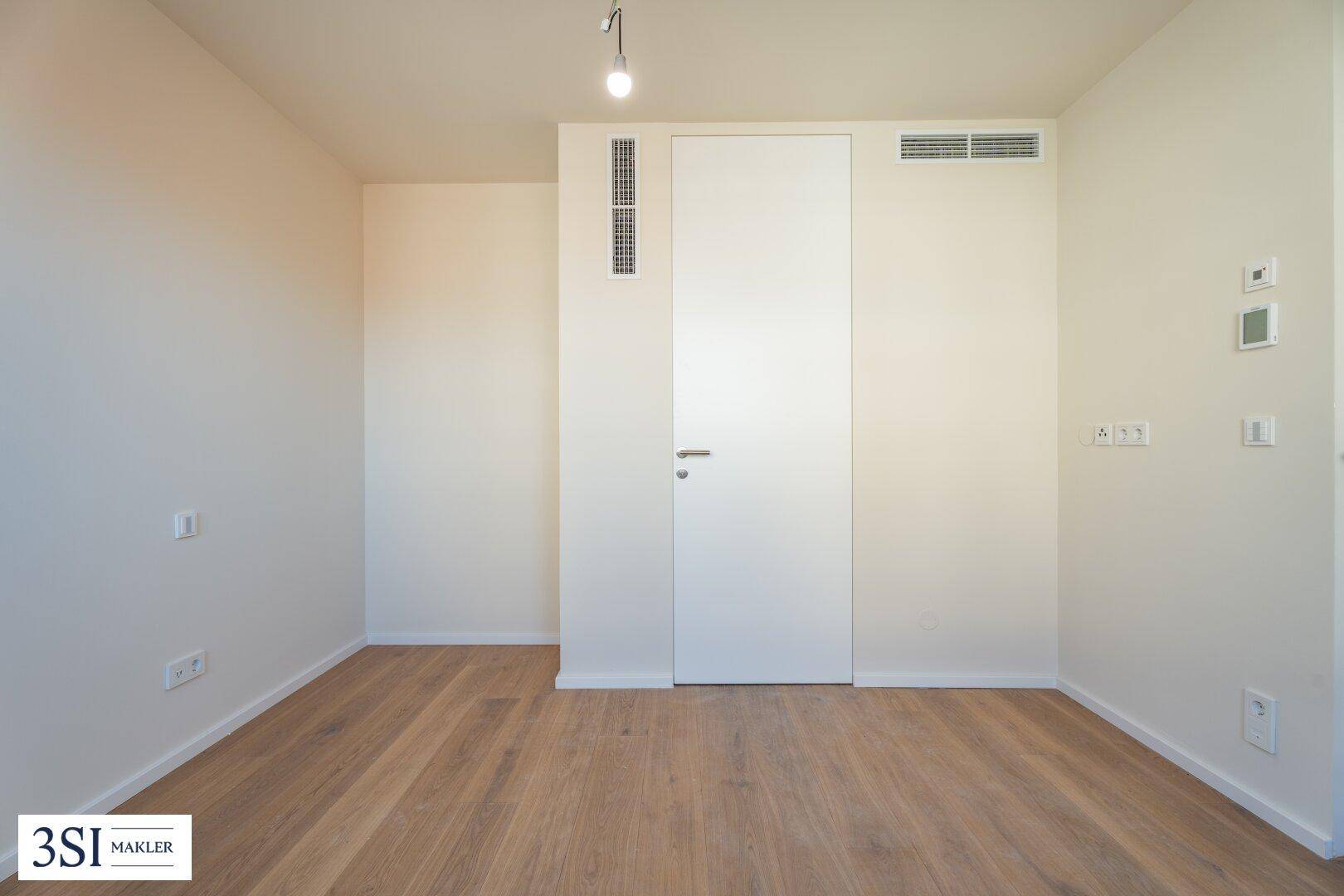 Heller Raum mit Holzboden, einer modernen Tür und Lüftungsschlitzen an der Decke.