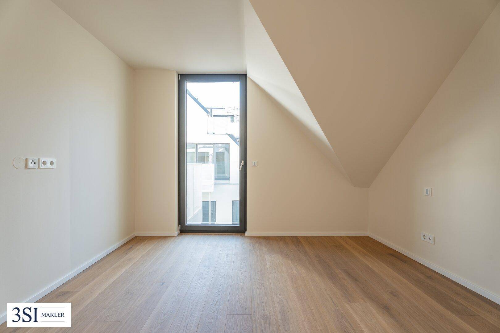 Dachgeschosszimmer mit Holzboden, schräger Decke und hohem Fenster, das viel Licht hereinlässt.