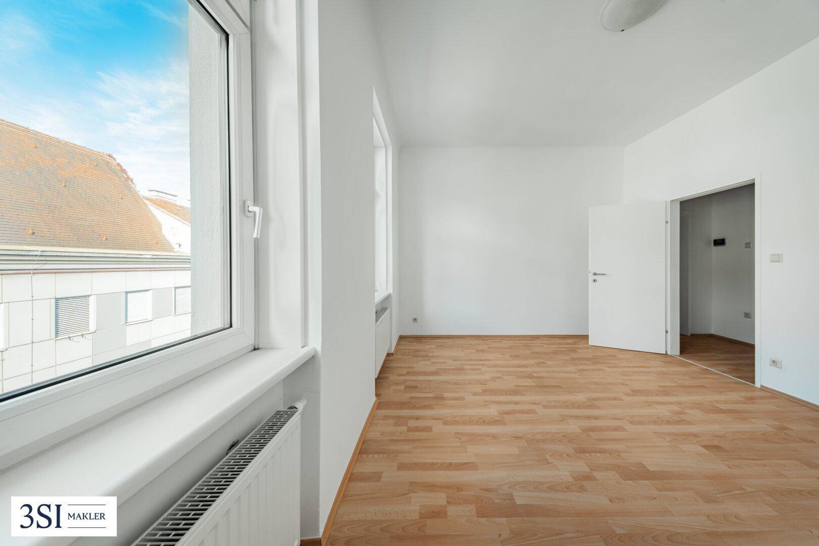 Lichtdurchfluteter Raum mit Parkettboden und großem Fenster, das einen Blick auf die Dächer bietet.