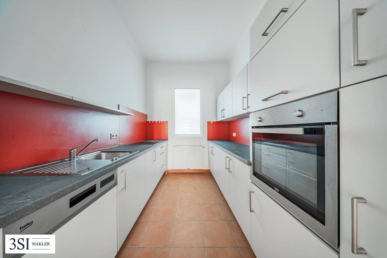 Moderne Einbauküche mit grauen Fronten, roter Akzentwand und Fenster für Tageslicht.