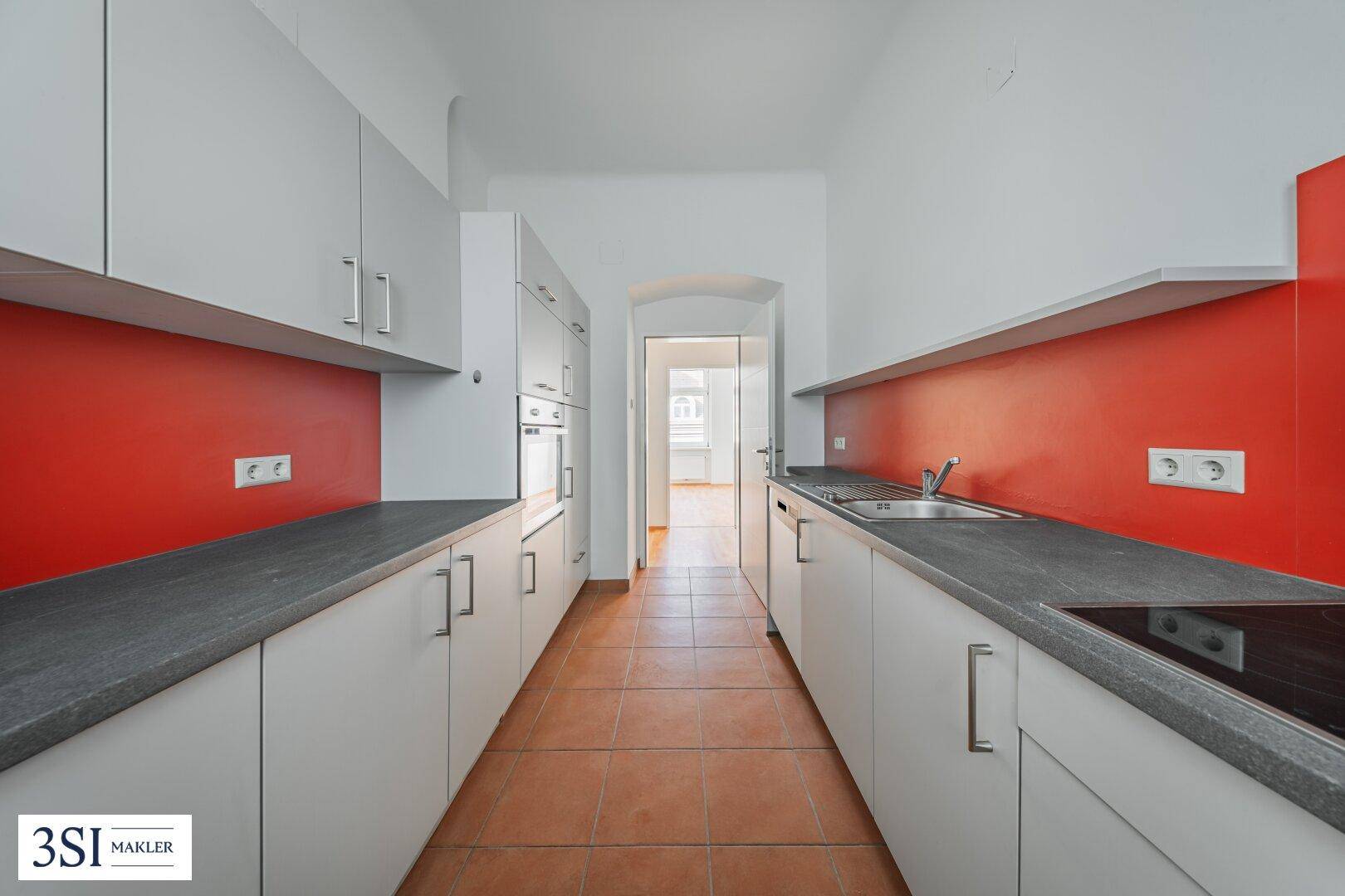 Funktionale Küche mit grauen Schränken, roter Rückwand und Terrakotta-Fliesenboden.