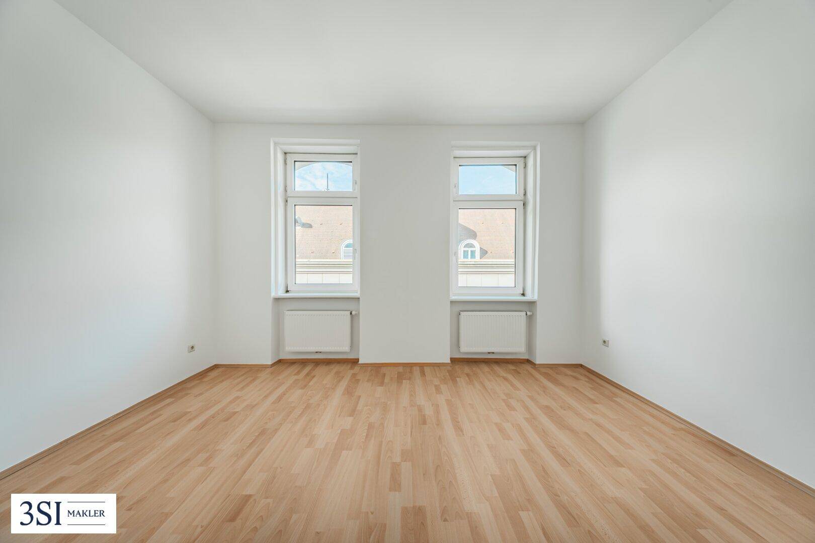 Helles, leeres Zimmer mit Parkettboden und zwei Fenstern, bereit zur individuellen Gestaltung.