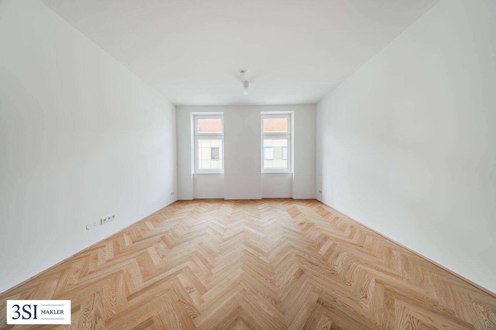 Helles, leeres Zimmer mit Fischgrätparkett und zwei Fenstern, bereit zur individuellen Gestaltung.