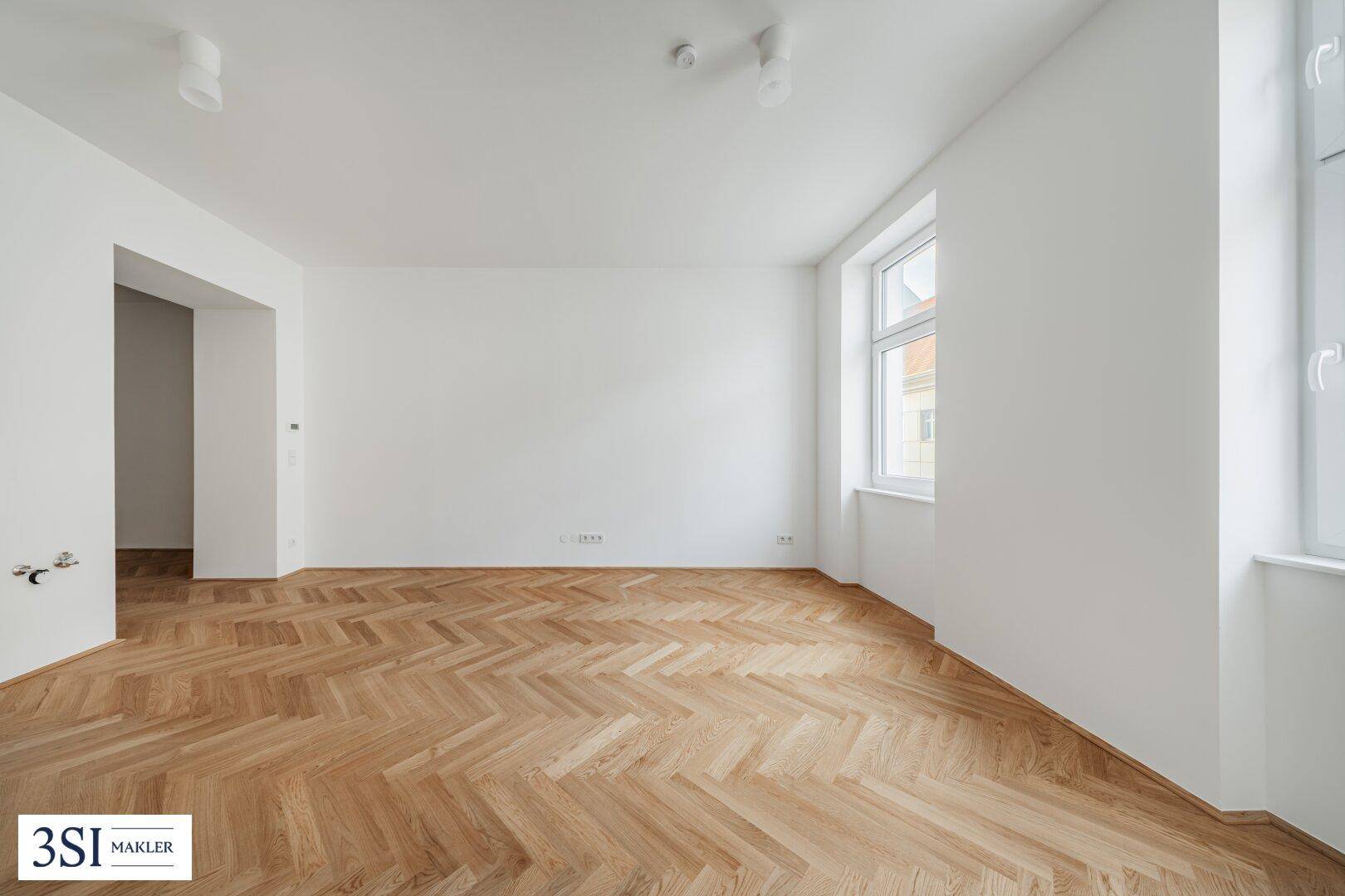 Geräumiges, helles Zimmer mit Fischgrätparkett und einem großen Fenster, ideal für Wohnzwecke.
