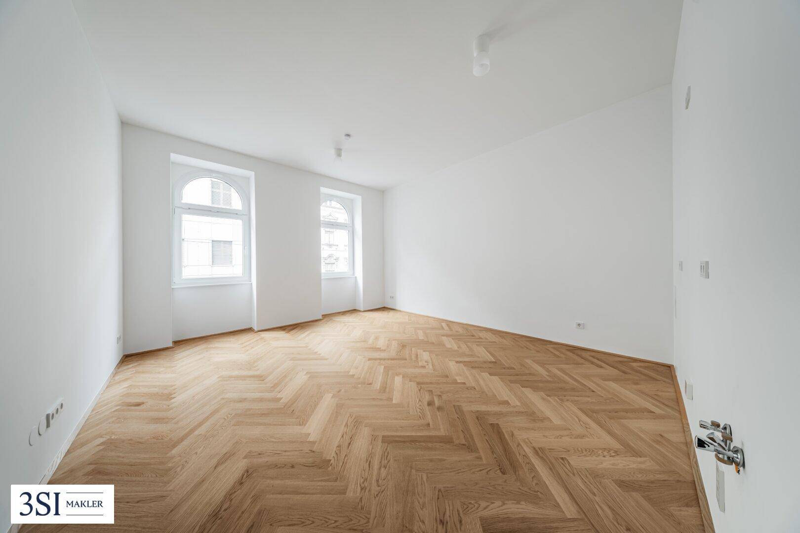 Geräumiger, heller Raum mit elegantem Fischgrätparkett und zwei hohen Rundbogenfenstern.