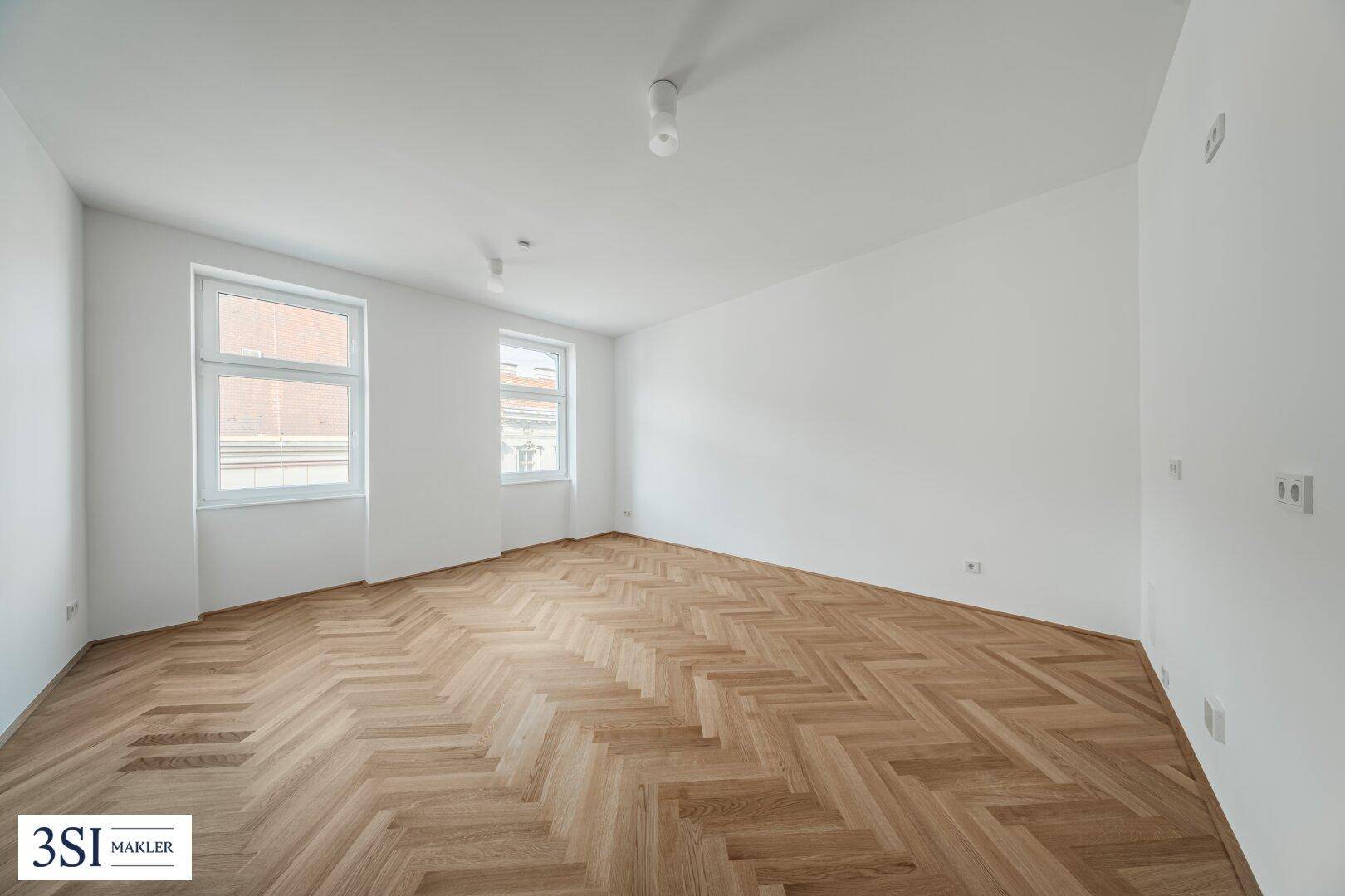 Geräumiges, helles Zimmer mit elegantem Fischgrätparkett und schlichten weißen Wänden.