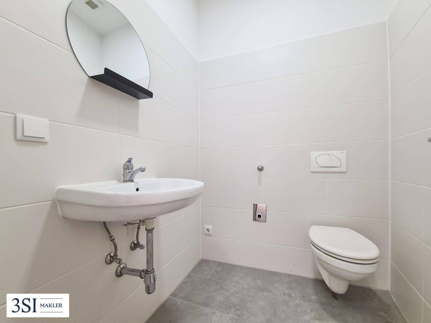 Modernes WC mit wandhängender Toilette, Waschbecken und Spiegel, sauber und hell gefliest.