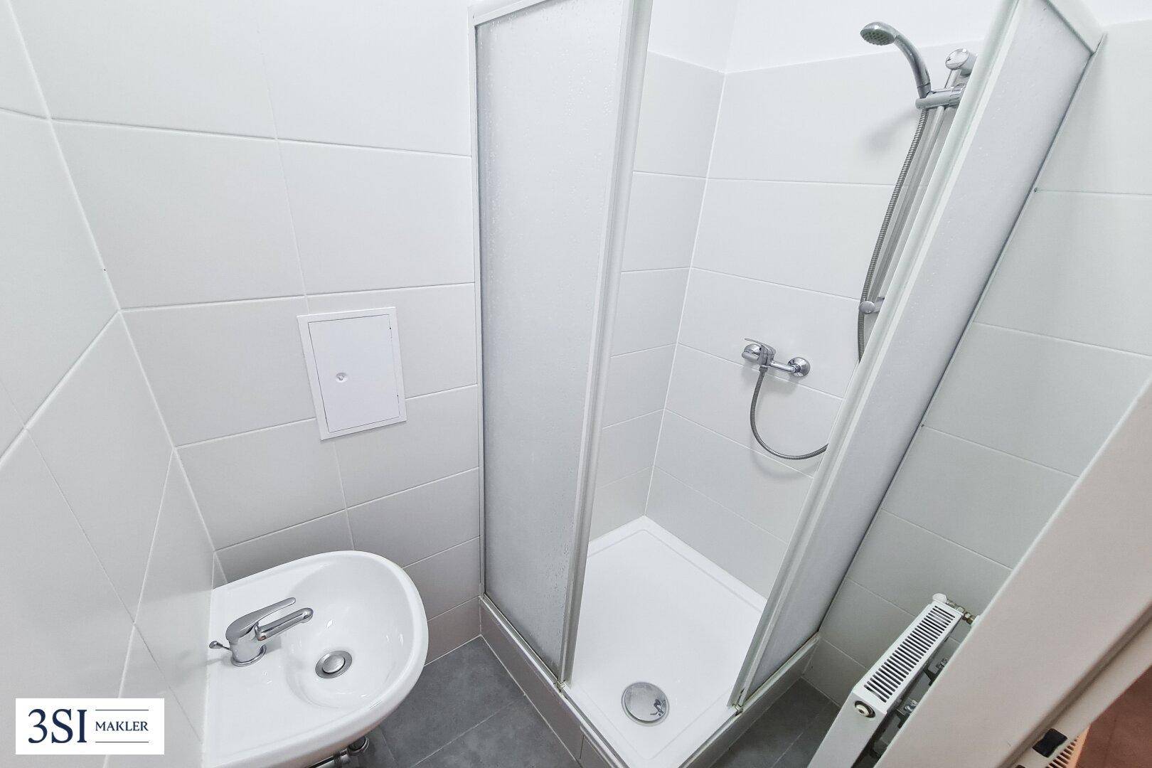 Kleines Badezimmer mit Duschkabine, Waschbecken und Heizkörper, hell gefliest und funktional.