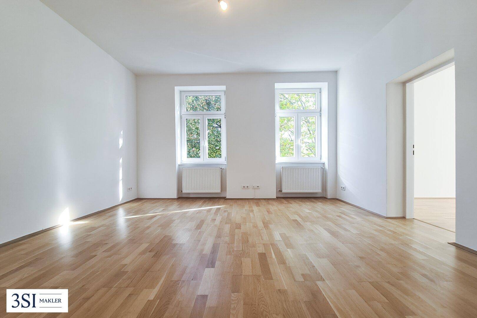 Geräumiges, unmöbliertes Wohnzimmer mit Holzboden und zwei großen Fenstern, bereit zur individuellen Gestaltung.