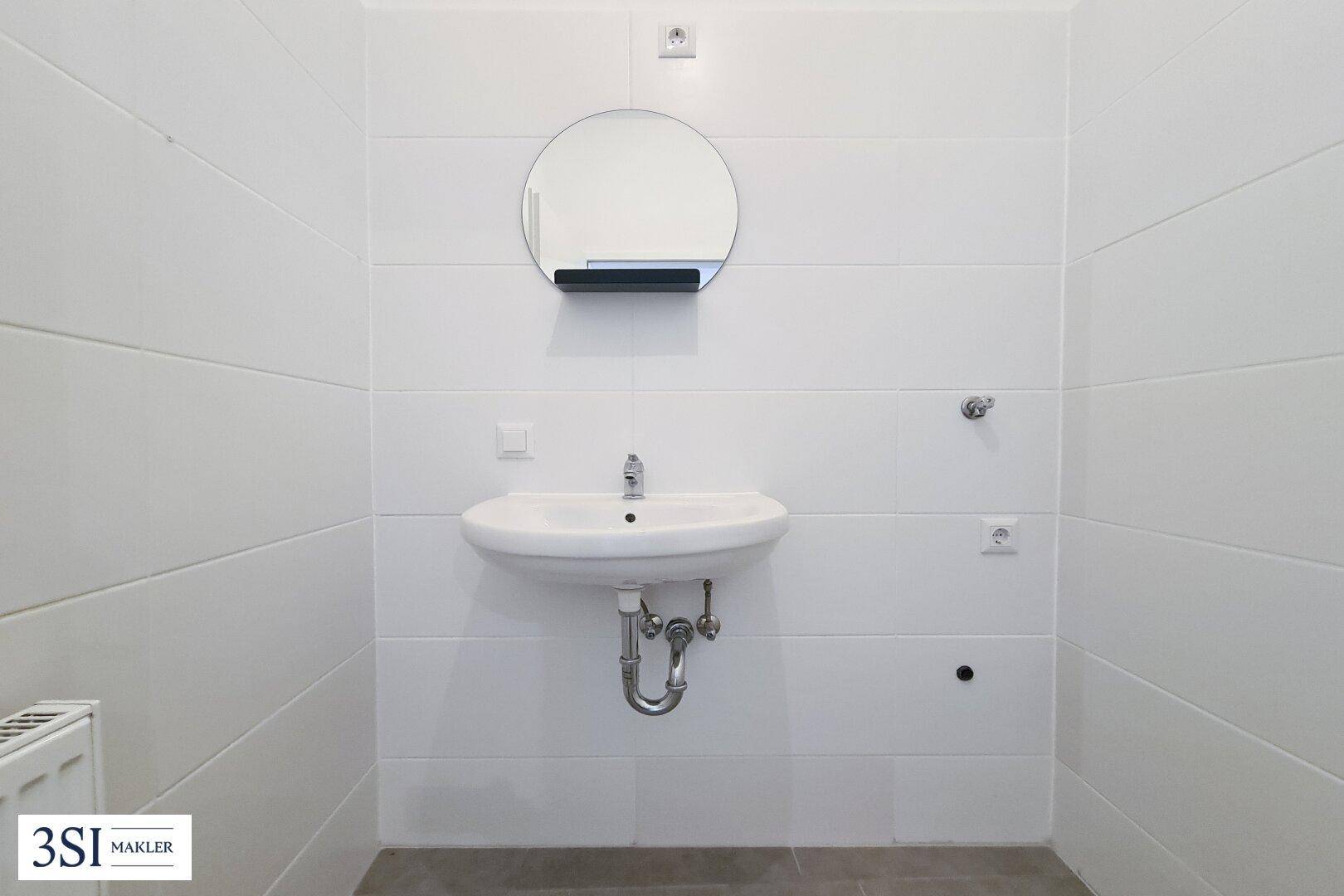 Modernes Badezimmer mit weißem Waschbecken, rundem Spiegel und hellen Fliesen.