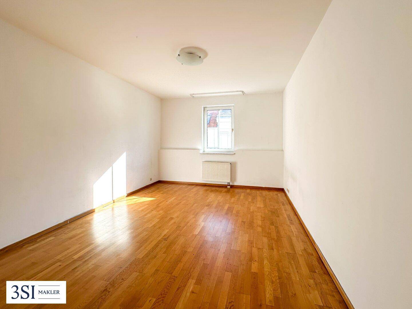 Heller, leerer Raum mit Holzboden und einem Fenster, das viel Sonnenlicht bietet.