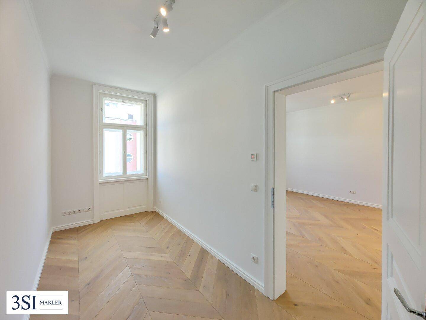 Lichtdurchfluteter Raum mit Fischgrätparkett und offener Verbindung zu einem weiteren Zimmer.