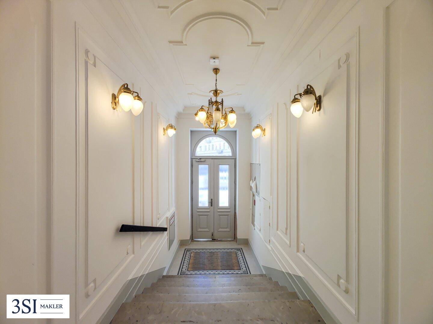 Elegantes Treppenhaus mit Stuckelementen, klassischer Beleuchtung und Doppeltür am Ende.