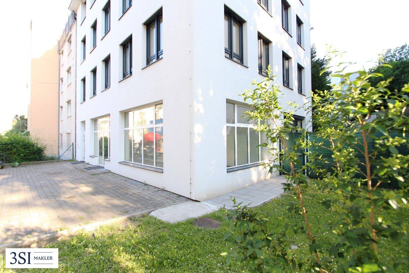 Moderne Gebäudefassade mit großen Fenstern im Erdgeschoss und einem kleinen Grünbereich.
