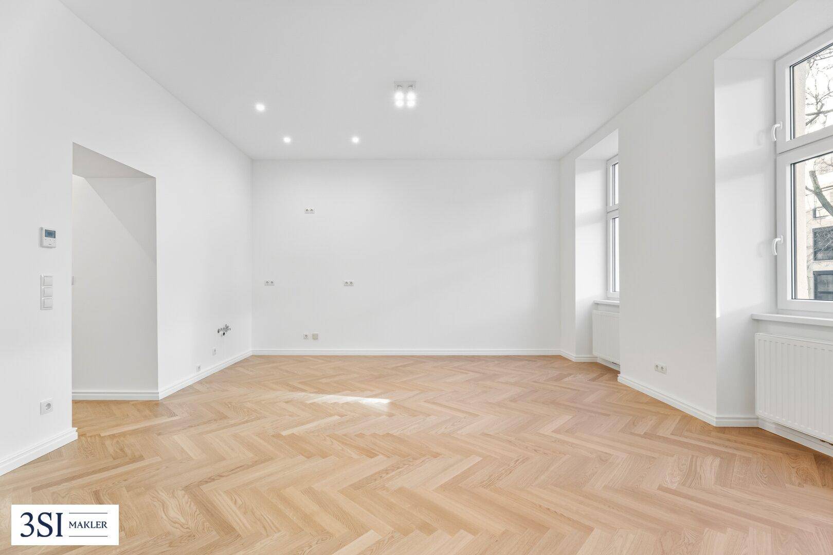 Weitläufiger, heller Raum mit Fischgrätparkett und großen Fenstern, bietet viel Tageslicht.