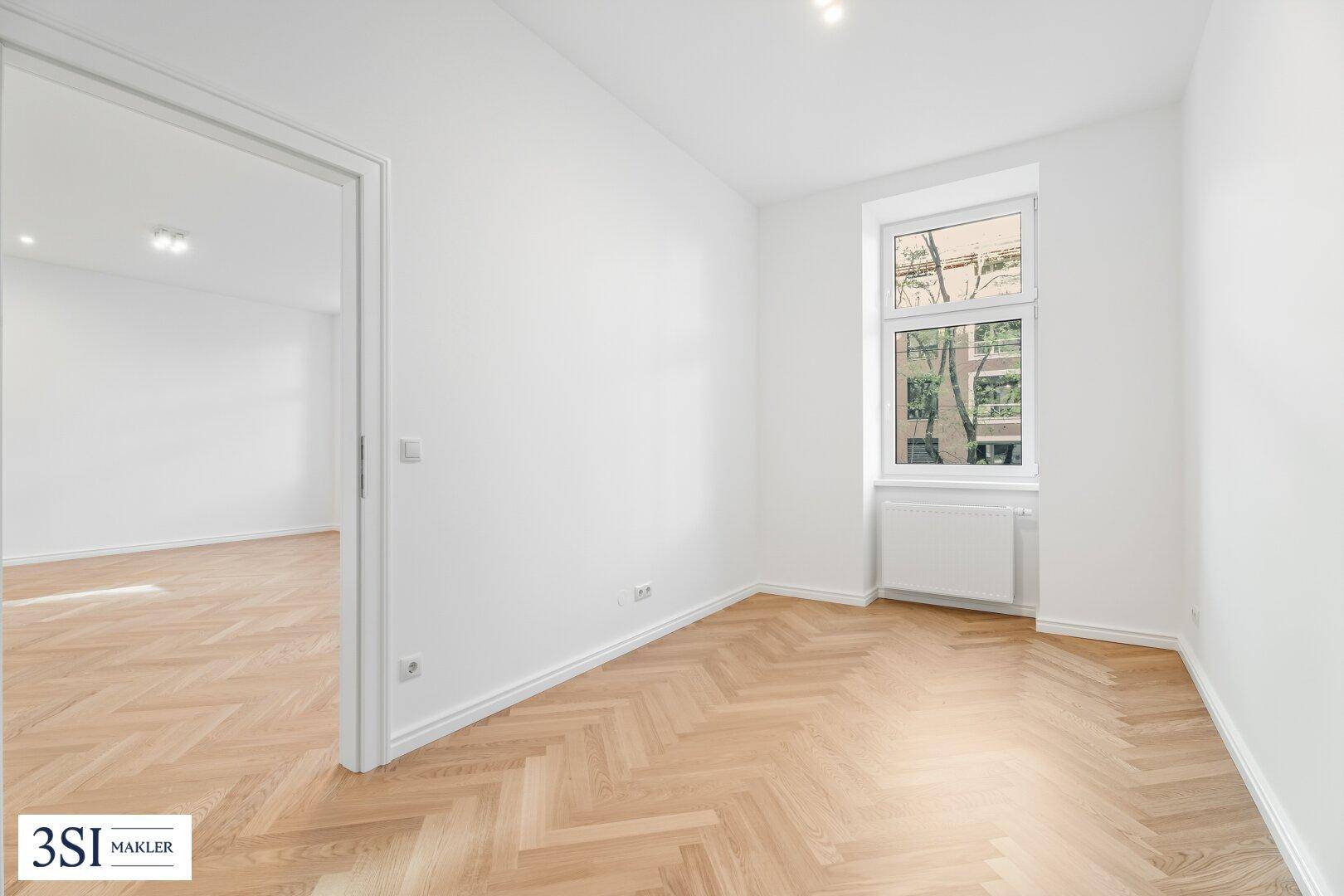 Helles Zimmer mit Fischgrätparkett und Fenster, bietet Zugang zu einem weiteren Raum.