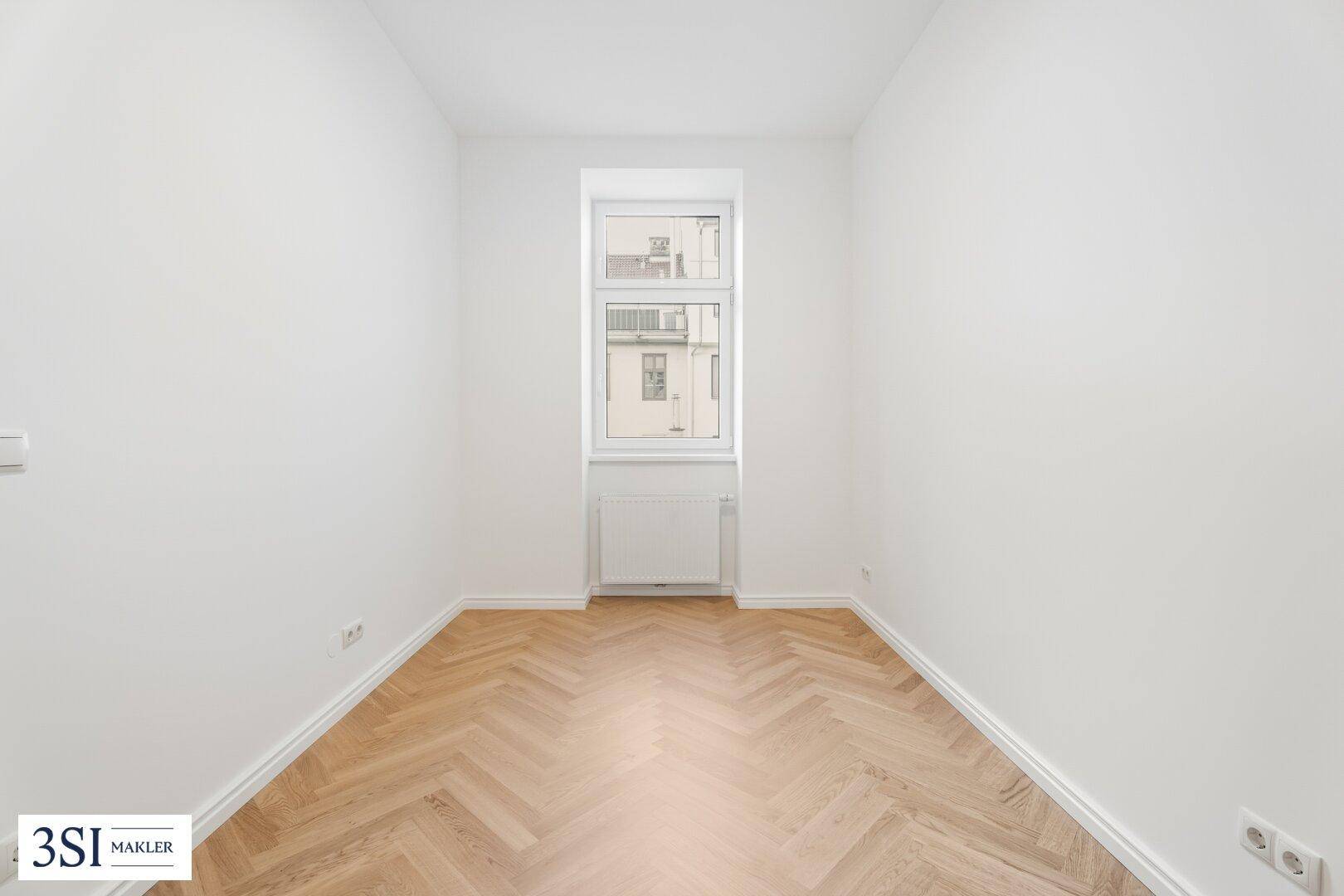 Ruhiges Zimmer mit Fischgrätparkett und einem einzelnen Fenster für natürliches Licht.