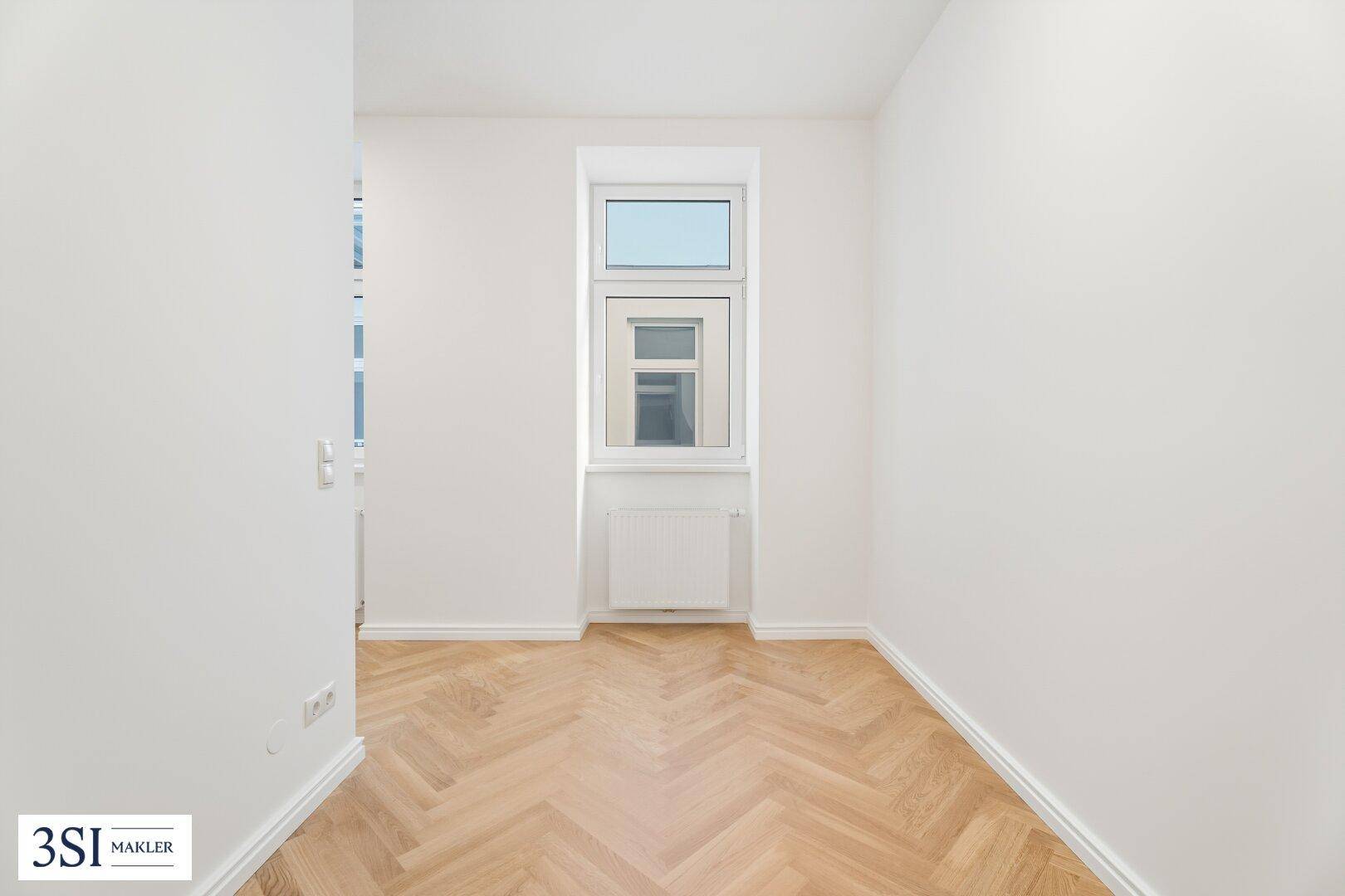 Schmaler Raum mit Fischgrätparkett und einem Fenster, ideal als Arbeitsbereich oder Flur.