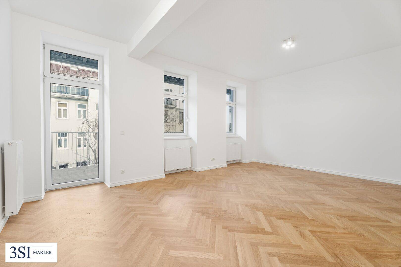 Geräumiges Zimmer mit hellem Fischgrätparkett und hohen Fenstern, die viel Tageslicht hereinlassen.