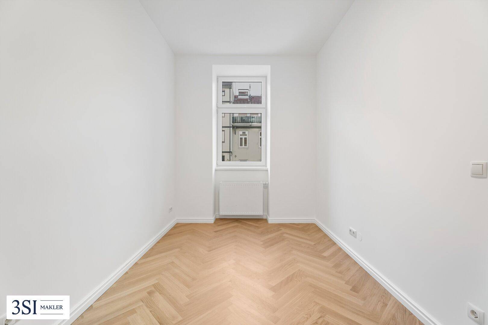 Helles Zimmer mit Fischgrätparkett und einem hohen Fenster, das für angenehmes Tageslicht sorgt.