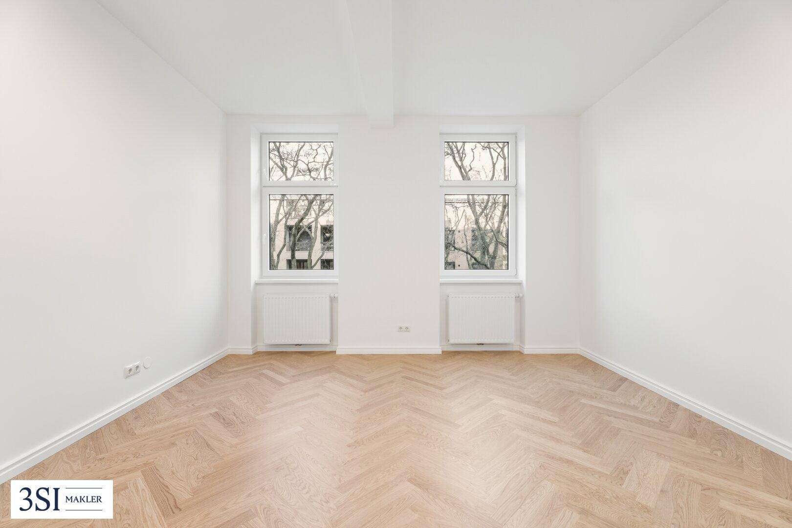 Leeres, helles Zimmer mit Fischgrätparkett und zwei hohen Fenstern.