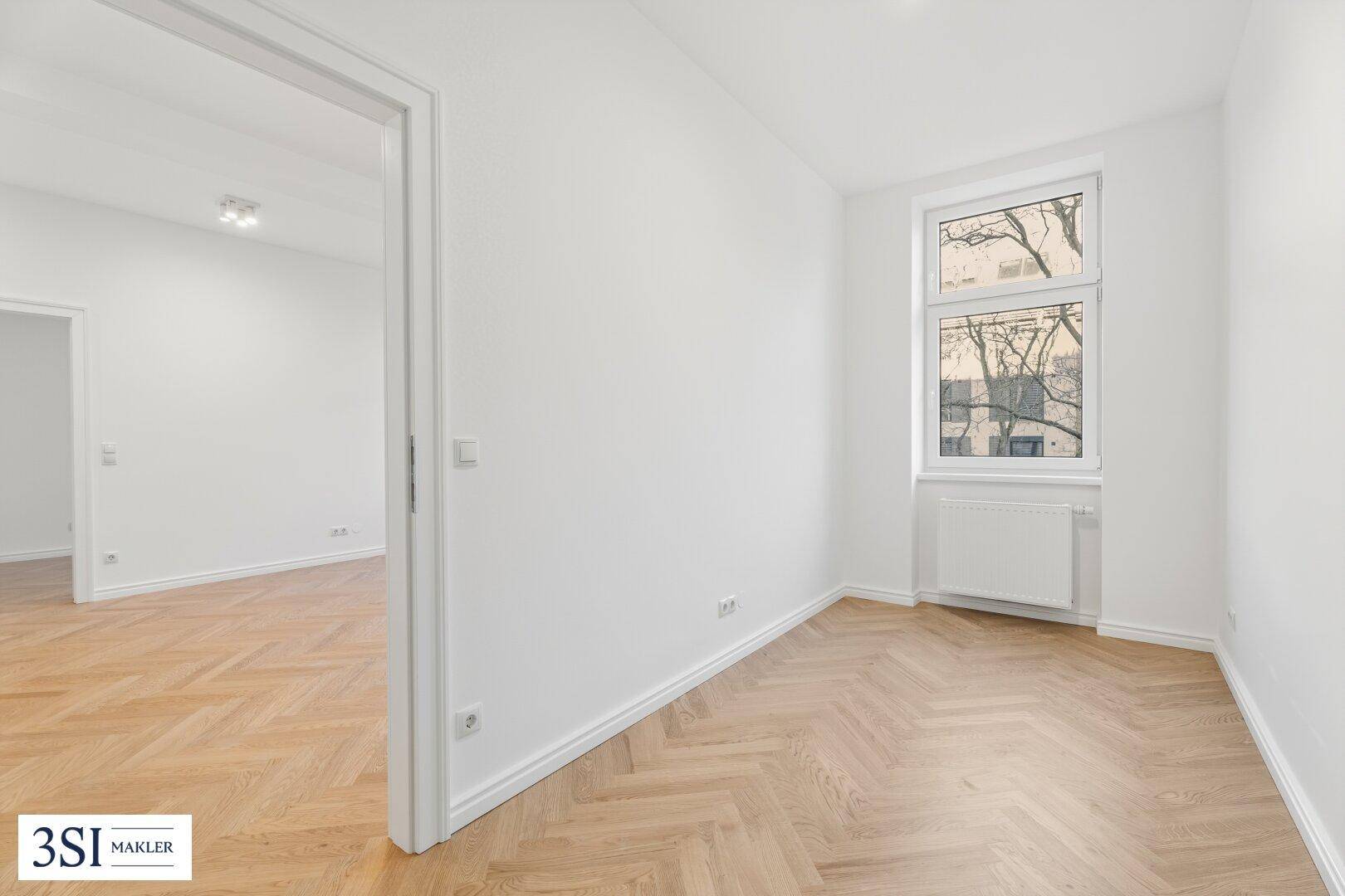 Helles Zimmer mit Fischgrätparkett und einer Tür, die zu weiteren Räumen führt.