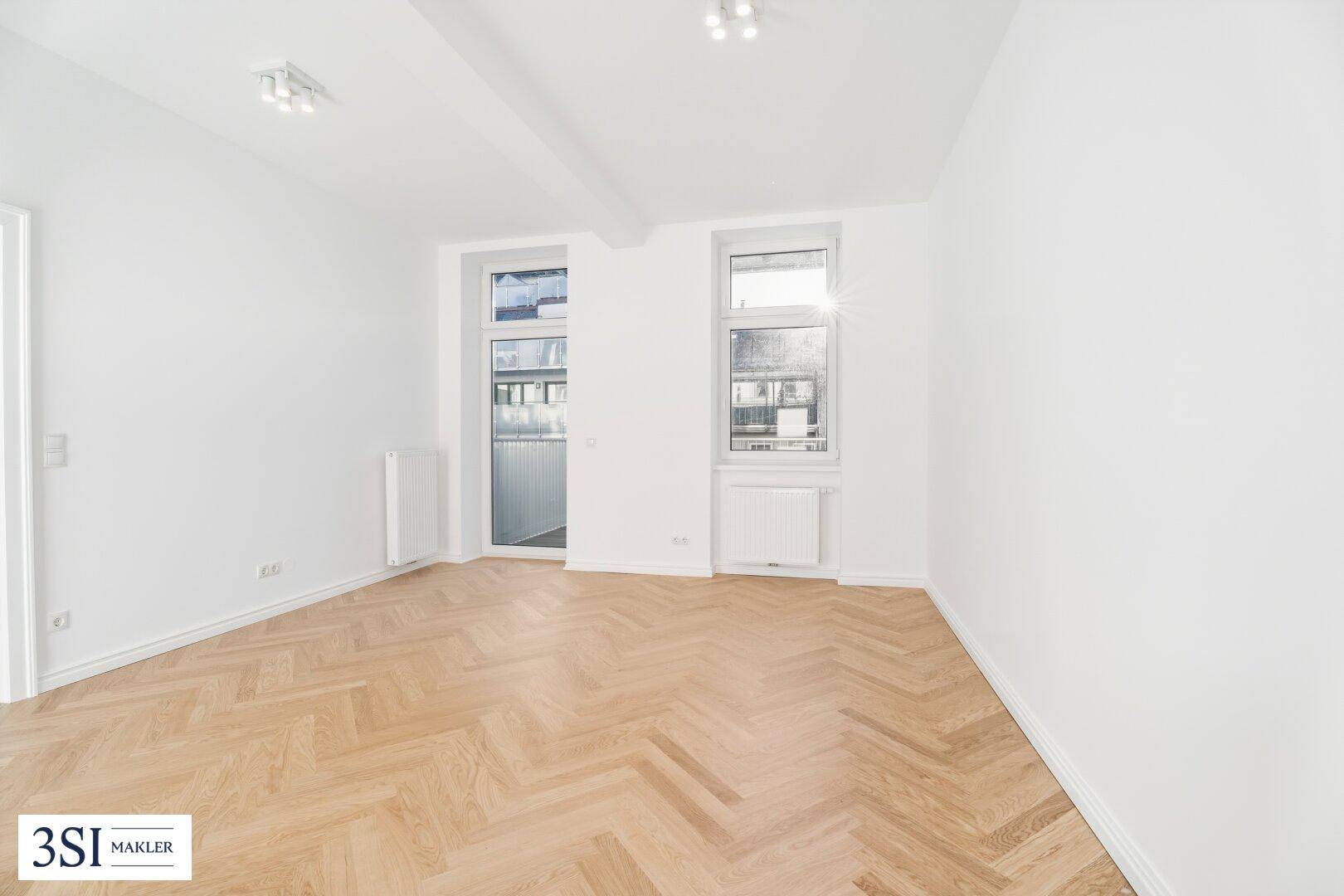 Helles, leeres Zimmer mit Fischgrätparkett und Zugang zu einem französischen Balkon.