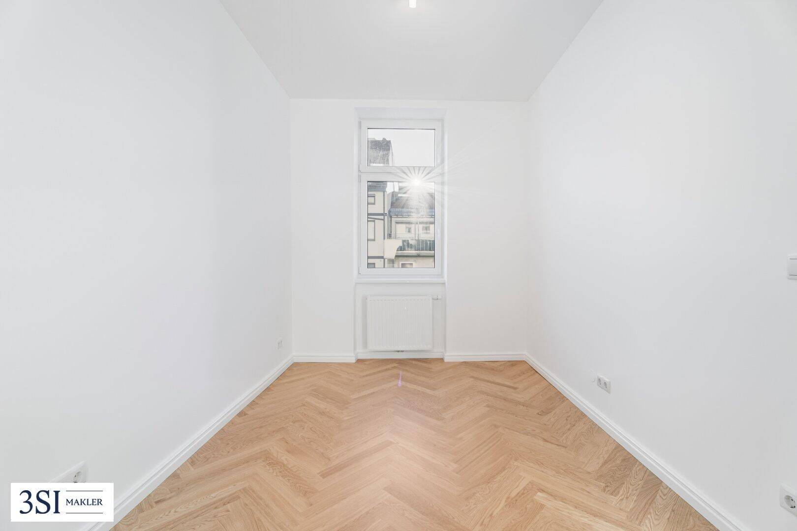Kleines, helles Zimmer mit Fischgrätparkett und einem Fenster, das viel Tageslicht hereinlässt.