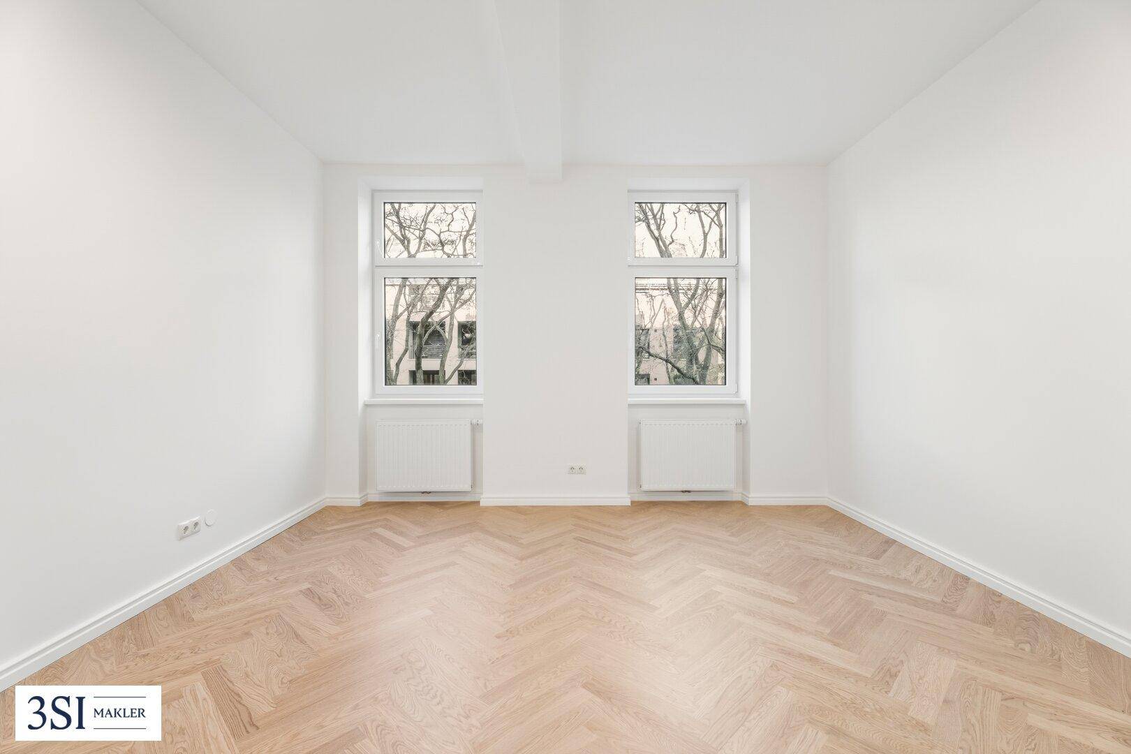 Geräumiges, helles Zimmer mit klassischem Fischgrätparkett und zwei großen Fenstern.