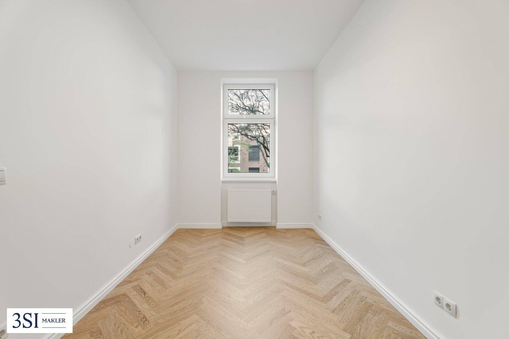 Schmaler, heller Raum mit Fischgrätparkett und einem Fenster, ideal als Schlaf- oder Arbeitszimmer.